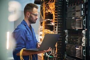 Silver colored laptop ,in, hands. Young man is working with internet equipment and wires in server room photo