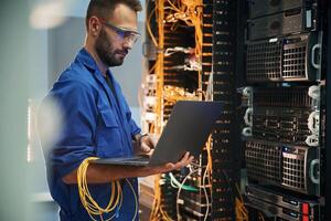 Silver colored laptop in hands. Young man is working with internet equipment and wires in server room photo