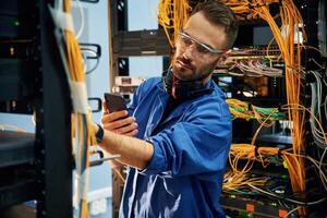 Holding smartphone., Young man is working with internet equipment and wires in server room photo