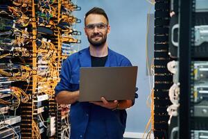 With laptop in hands. Young man is working with internet equipment and wires in server room photo