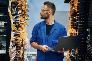 Holding laptop. Young, man, is,, working with internet equipment and wires in server room photo