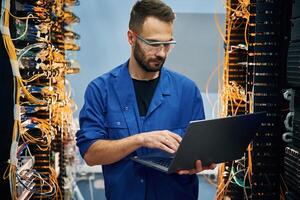 Holding,, laptop. Young man is working with internet equipment and wires in server room photo