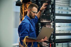 Using, the laptop. Young ,man is working with internet equipment and wires in server room photo