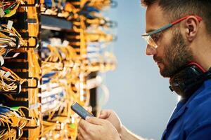 Holding ,smartphone.. Young man is working with internet equipment and wires in server room photo