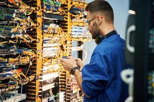 Holding ,smartphone. Young man, is working with internet equipment and wires in server room photo