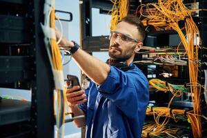 Holding smartphone... Young man is working with internet equipment and wires in server room photo