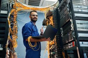 Silver colored laptop in hands.,. Young man is working with internet equipment and wires in server room photo