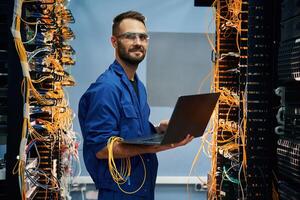 With laptop ,in hands. Young man is working with internet equipment and wires in server room photo