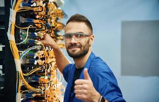 Fixed, with thumbs up.,. Young man is working with internet equipment and wires in server room photo