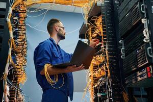 Silver, colored laptop in hands. Young man is working with internet equipment and wires in server room photo