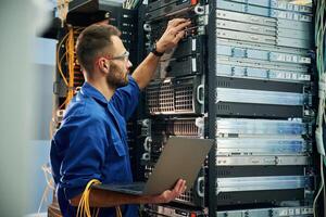 Using the laptop. Young man, is ,working with internet equipment and wires in server room photo