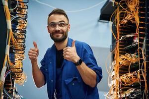 Fixed, with thumbs up,. Young man is working with internet equipment and wires in server room photo