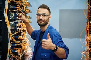 Fixed,, with thumbs up. Young man is working with internet equipment and wires in server room photo