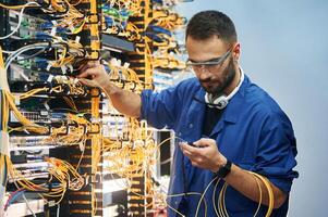 Checking signal, by ,device. Young man is working with internet equipment and wires in server room photo