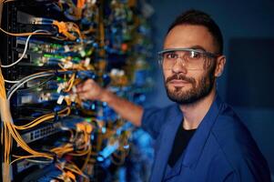 Looking to, the camera. Young man is working with internet equipment and wires in server room photo
