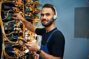 Headphones ,and smartphone.. Young man is working with internet equipment and wires in server room photo