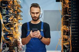 Headphones and, smartphone. Young man is working with internet equipment and wires in server room photo