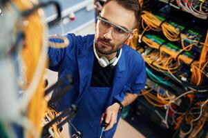 In blue, colored uniform. Young man is working with internet equipment and wires in server room photo