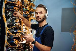 Headphones, and smartphone. Young man is working with internet equipment and wires in server room photo