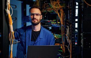 Looking, to the camera. Young man is working with internet equipment and wires in server room photo