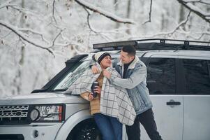 Man is covering ,woman with, warm blanket. Beautiful couple standing near the car in the winter forest photo