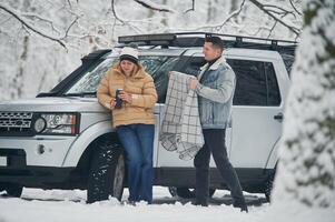 Man, is covering woman with warm blanket. Beautiful couple standing near the car in the winter forest photo