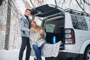 Woman is sitting ,in the trunk. Beautiful couple standing near the car in the winter forest photo