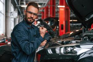 Looking at the camera., Car repairman is in the garage with automobile photo