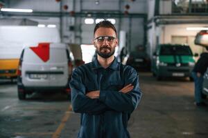 With arms crossed,,, standing. Car repairman is in the garage with automobile photo