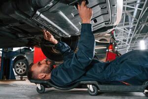 Lying down, on ,the ground. Car repairman is in the garage with automobile photo