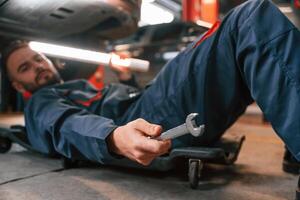 Lying down ,on the ground. Car repairman is in the garage with automobile photo