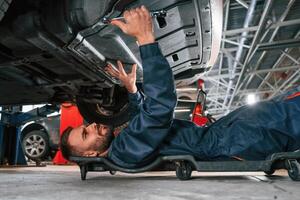 On the ground with, tool. Car repairman is in the garage with automobile photo