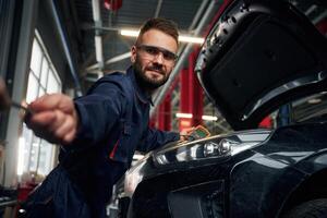 Looking ,at the camera. Car repairman is in the garage with automobile photo