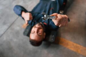 On the ground, with tool. Car repairman is in the garage with automobile photo