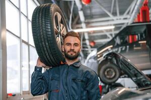 Holding, a, tire. Car repairman is in the garage with automobile photo