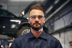 Looking, at the camera. Car repairman is in the garage with automobile photo