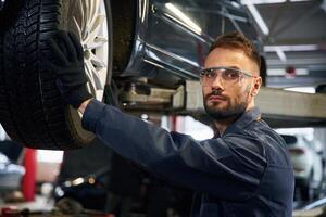 Looking at, the camera. Car repairman is in the garage with automobile photo