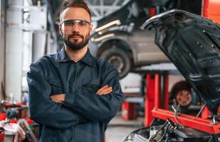 With arms ,crossed, standing. Car repairman is in the garage with automobile photo