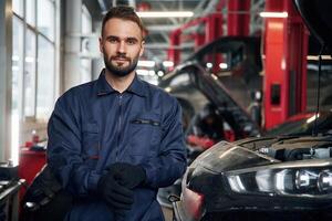 Wearing the ,gloves, standing. Car repairman is in the garage with automobile photo
