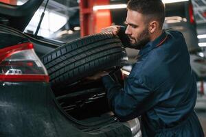 Holding ,a tire. Car repairman is in the garage with automobile photo