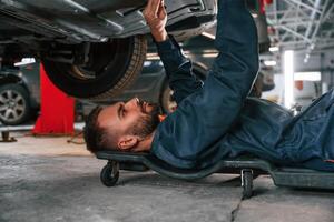 Lying,, down on the ground. Car repairman is in the garage with automobile photo