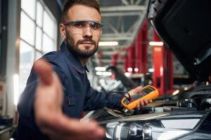 Yellow, colored device for measuring electricity is in hands. Car repairman is in the garage with automobile photo