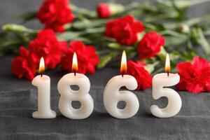 Burning candles forming the number 1865 with red carnations in the background photo