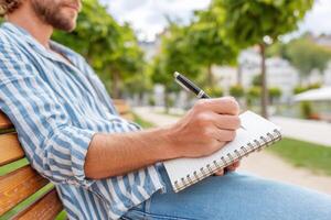 Man writing notes in notebook sitting on bench in city park photo
