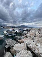 Boats anchored at harbor with rocky shoreline under cloudy sky in evening light photo