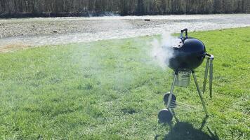 Round charcoal grill smoking on grassy riverside during outdoor barbecue, portable kettle grill in use for cooking with smoke rising under sunny spring weather video