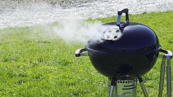 Round kettle grill with built-in thermometer smoking on riverside lawn, black charcoal barbecue in use for outdoor cooking under bright spring sunlight video