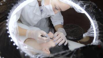 Beautician performing permanent eyeliner procedure using tattoo machine, client lying under LED ring light while technician in mask and gloves works with precision video