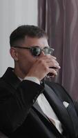 Stylish man in a black suit and sunglasses, drinking in a relaxed indoor setting . Vertical video