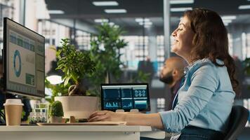 Cheerful employee at office desk having fun with coworkers while editing documents on computer. Happy worker sharing jokes with colleagues while analyzing data charts on PC, camera A video